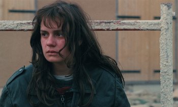 Movie still from “Vagabond” (1985), directed by Agnès Varda – A woman with long brown hair is standing in front of a pole; Close Up shot, Over the shoulder angle