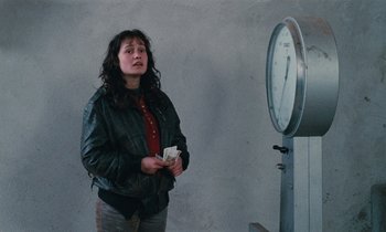 Movie still from “Vagabond” (1985), directed by Agnès Varda – A woman standing in front of a clock holding a piece of paper; Medium shot, Over the shoulder angle