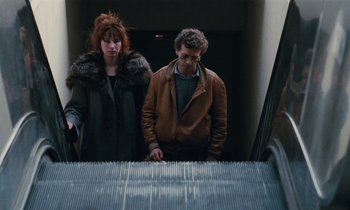 Movie still from “Vagabond” (1985), directed by Agnès Varda – A man and a woman standing next to an escalator; Medium shot, High angle