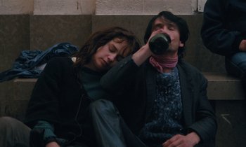 Movie still from “Vagabond” (1985), directed by Agnès Varda – A man and a woman sitting next to each other drinking; Medium shot, High angle
