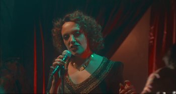 Movie still from “Velvet Goldmine” (1998), directed by Todd Haynes – A woman with curly hair is holding a microphone; Medium shot, Low angle