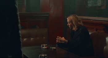 Movie still from “Velvet Goldmine” (1998), directed by Todd Haynes – A woman sitting at a table with a cigarette in her hand; Medium shot, Over the shoulder angle
