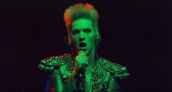 Movie still from “Velvet Goldmine” (1998), directed by Todd Haynes – A man in a green jacket singing into a microphone; Close Up shot, Low angle