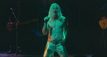 Movie still from “Velvet Goldmine” (1998), directed by Todd Haynes – A man with long blond hair is holding a microphone and singing; Medium shot, Low angle