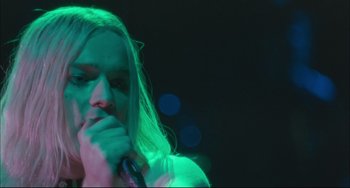 Movie still from “Velvet Goldmine” (1998), directed by Todd Haynes – A man with long blonde hair is holding a microphone; Close Up shot, Low angle