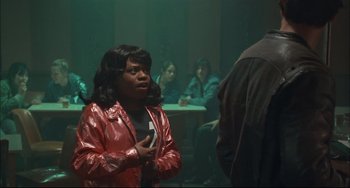 Movie still from “Velvet Goldmine” (1998), directed by Todd Haynes – A woman in a red jacket talking to a group of people in a room; Medium shot, Over the shoulder angle