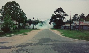 Movie still from “Vernon, Florida” (1981), directed by Errol Morris – Smoke billows out of the street as a car drives down the road; Extreme Wide shot, High angle