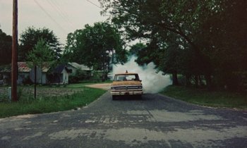 Movie still from “Vernon, Florida” (1981), directed by Errol Morris – An old car is driving down the street with smoke coming out of the hood; Extreme Wide shot, High angle