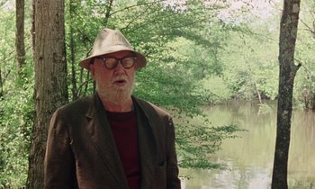 Movie still from “Vernon, Florida” (1981), directed by Errol Morris – An older man wearing a hat standing next to a body of water; Medium shot, Over the shoulder angle