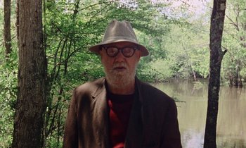 Movie still from “Vernon, Florida” (1981), directed by Errol Morris – An older man wearing a hat standing in front of a body of water; Medium shot, Low angle