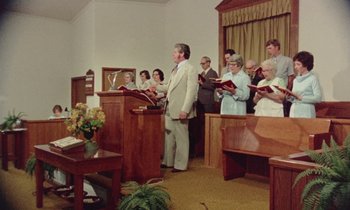 Movie still from “Vernon, Florida” (1981), directed by Errol Morris – A group of people standing in a room with microphones; Wide shot, Low angle