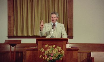 Movie still from “Vernon, Florida” (1981), directed by Errol Morris – A man in a suit giving a speech at a podium; Medium shot, High angle
