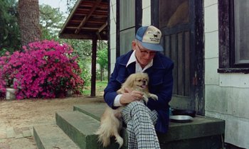 Movie still from “Vernon, Florida” (1981), directed by Errol Morris – An older man sitting on the steps holding a dog; Medium shot, High angle