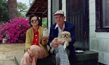 Movie still from “Vernon, Florida” (1981), directed by Errol Morris – An older man and woman sitting next to each other holding a dog; Medium shot, High angle