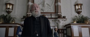 Movie still from “Victoria & Abdul” (2017), directed by Stephen Frears – An old man with a long beard standing in front of a statue of an angel; Medium shot, Low angle