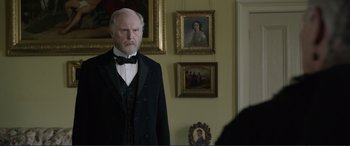 Movie still from “Victoria & Abdul” (2017), directed by Stephen Frears – An older man in a suit and bow tie in front of a painting; Medium shot, Over the shoulder angle