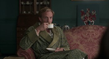 Movie still from “Vita & Virginia” (2018), directed by Chanya Button – A man sitting on top of a couch drinking a cup of coffee; Medium shot, High angle