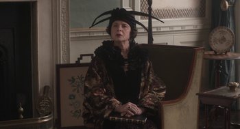 Movie still from “Vita & Virginia” (2018), directed by Chanya Button – An older woman sitting on top of a chair wearing a hat; Medium shot, Low angle