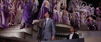 Movie still from “Viva Las Vegas” (1964), directed by George Sidney – A man in a gray suit standing in front of a stage with showgirls; Wide shot, Low angle