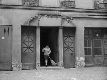 Movie still from “My Life to Live” (1962), directed by Jean-Luc Godard – A man holding an object in a doorway of a building; Wide shot, High angle