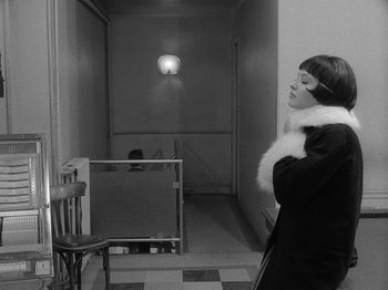 Movie still from “My Life to Live” (1962), directed by Jean-Luc Godard – A black and white photo of a woman standing in front of a chair; Medium shot, Low angle