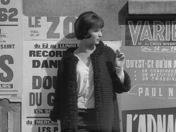 Movie still from “My Life to Live” (1962), directed by Jean-Luc Godard – A woman smoking a cigarette in front of a wall of posters; Medium shot, High angle