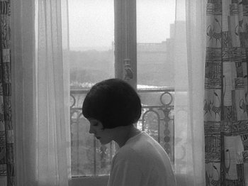 Movie still from “My Life to Live” (1962), directed by Jean-Luc Godard – A woman standing in front of a window with curtains; Medium shot, High angle