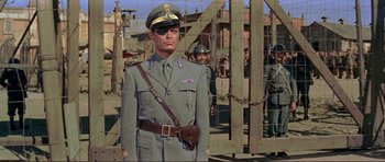 Movie still from “Von Ryan's Express” (1965), directed by Mark Robson – A man in a military uniform standing in front of a fence; Medium shot, Low angle