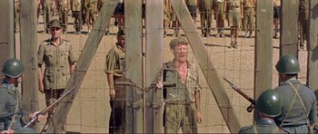 Movie still from “Von Ryan's Express” (1965), directed by Mark Robson – A man chained to a metal fence in front of other men; Wide shot, Low angle