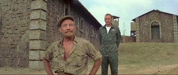 Movie still from “Von Ryan's Express” (1965), directed by Mark Robson – Two men standing next to each other in front of a brick building; Medium shot, Over the shoulder angle