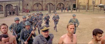 Movie still from “Von Ryan's Express” (1965), directed by Mark Robson – A group of men in uniform marching on a dirt field; Wide shot, Over the shoulder angle