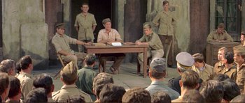 Movie still from “Von Ryan's Express” (1965), directed by Mark Robson – A group of men sitting at a table in front of an audience; Wide shot, High angle