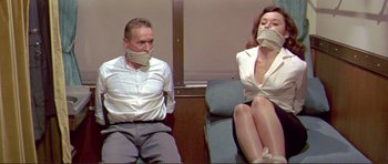 Movie still from “Von Ryan's Express” (1965), directed by Mark Robson – A man and a woman sitting on a couch with their mouths taped shut; Medium shot, Low angle