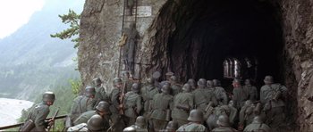 Movie still from “Von Ryan's Express” (1965), directed by Mark Robson – A group of men in uniform standing in front of an entrance to a cave; Wide shot, High angle