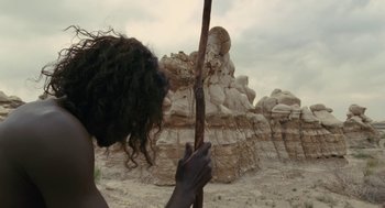 Movie still from “Voyage of Time” (2016), directed by Terrence Malick – A person holding a wooden stick in front of a rock formation; Medium shot, Low angle