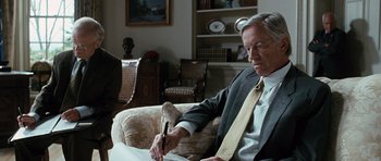 Movie still from “W.” (2008), directed by Oliver Stone – An older man sitting in a chair writing on a piece of paper; Medium shot, Over the shoulder angle