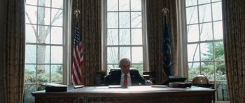 Movie still from “W.” (2008), directed by Oliver Stone – A man sitting at a desk in front of two american flags; Medium shot, Low angle
