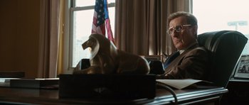 Movie still from “W.” (2008), directed by Oliver Stone – A man sitting in front of an american flag; Medium shot, Low angle