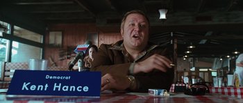 Movie still from “W.” (2008), directed by Oliver Stone – A man sitting in front of a microphone at a press conference; Medium shot, High angle