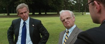 Movie still from “W.” (2008), directed by Oliver Stone – A man in a suit and tie standing next to another man in a suit and tie; Medium shot, Over the shoulder angle