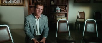 Movie still from “W.” (2008), directed by Oliver Stone – A man sitting in front of a chair in front of a book case; Medium shot, Over the shoulder angle