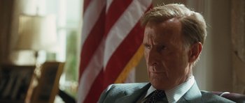 Movie still from “W.” (2008), directed by Oliver Stone – A man in a suit and tie in front of an american flag; Close Up shot, Low angle
