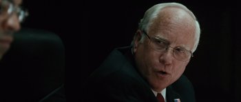 Movie still from “W.” (2008), directed by Oliver Stone – An older man wearing glasses and a suit and red tie; Close Up shot, Over the shoulder angle