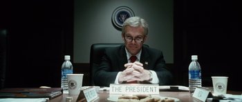 Movie still from “W.” (2008), directed by Oliver Stone – A man sitting in front of a table with cookies on top of it; Medium shot, Over the shoulder angle