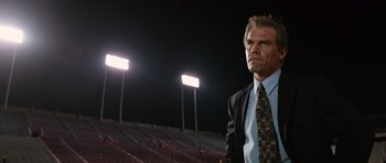 Movie still from “W.” (2008), directed by Oliver Stone – A man in a suit and tie standing in front of bleachers at night; Medium shot, Low angle