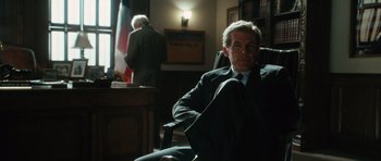 Movie still from “W.” (2008), directed by Oliver Stone – A man in a suit sitting in a chair in front of a desk; Medium shot, Low angle