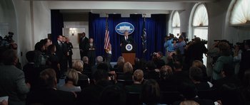 Movie still from “W.” (2008), directed by Oliver Stone – A man is speaking at a podium in front of an audience; Wide shot, High angle