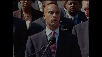 Movie still from “Waiting for Superman” (2010), directed by Davis Guggenheim – A man in a suit and tie standing in front of microphones; Medium shot, Low angle