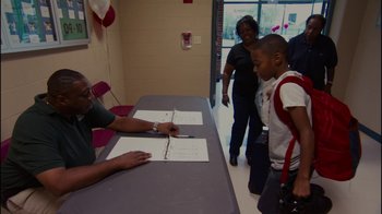 Movie still from “Waiting for Superman” (2010), directed by Davis Guggenheim – A man sitting at a table signing a book for a young boy; Medium shot, Over the shoulder angle