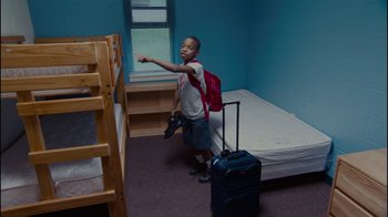 Movie still from “Waiting for Superman” (2010), directed by Davis Guggenheim – A young boy standing in front of a bed with a suitcase; Wide shot, High angle